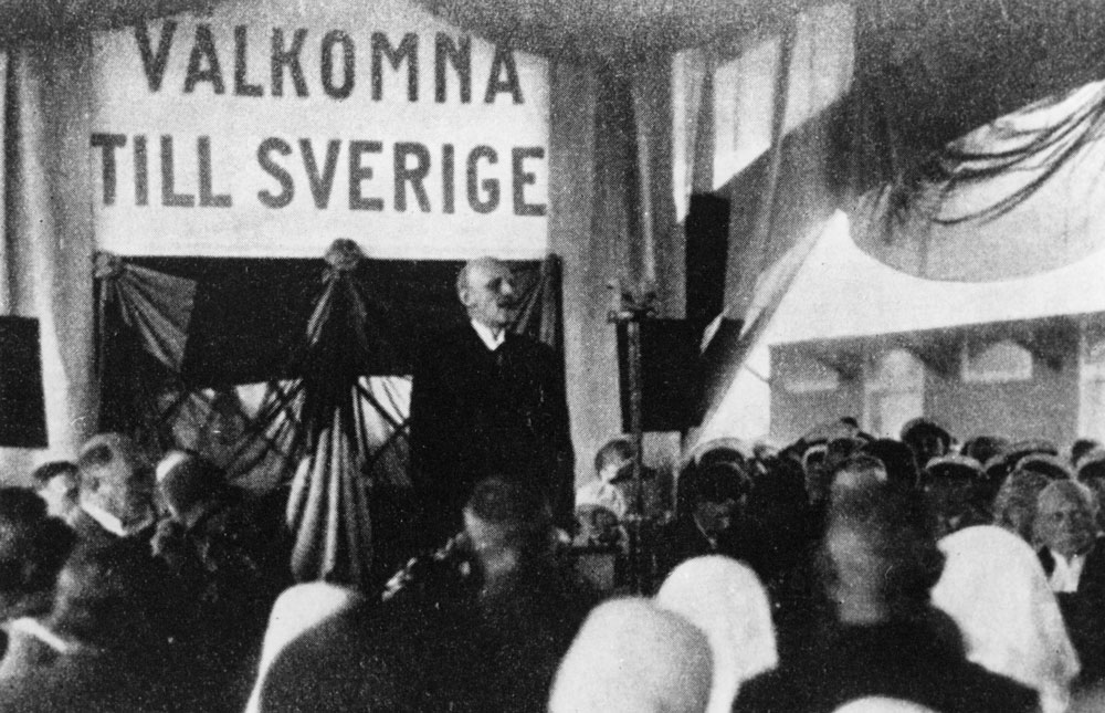 Man som håller tal i talarstol, i bakgrunden banderoll med texten "Välkommen till Sverige". 