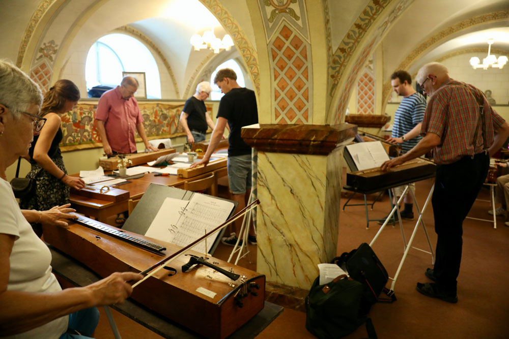 Sju personer står vid psalmodikon i ett rum och spelar.