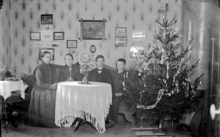 svartvitt fotografi med två vuxna och två barn som sitter i en soffa bredvid en klädd julgran  i ett vardagsliknande rum