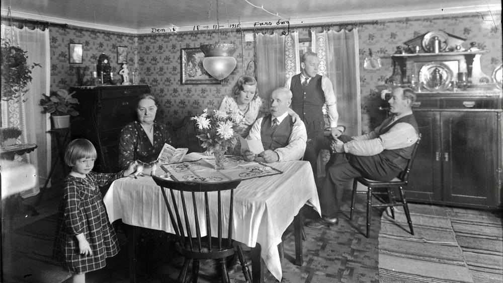 svartvitt fotografi med ett äldre par som sitter vid ett bord i ett vardagsrum. Runtomkring dem står och sitter tre yngre personer och ett barn
