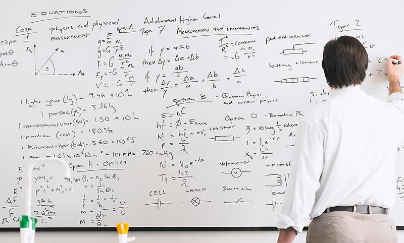En man står framför en whiteboard och skriver ekvationer, och enstaka ord. Orden är på engelska.