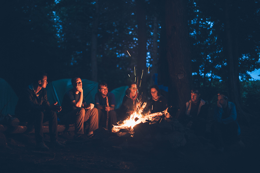 En grupp vuxna människor sitter i skogen runt en lägereld. Det är sen kväll, och mörkt ute.