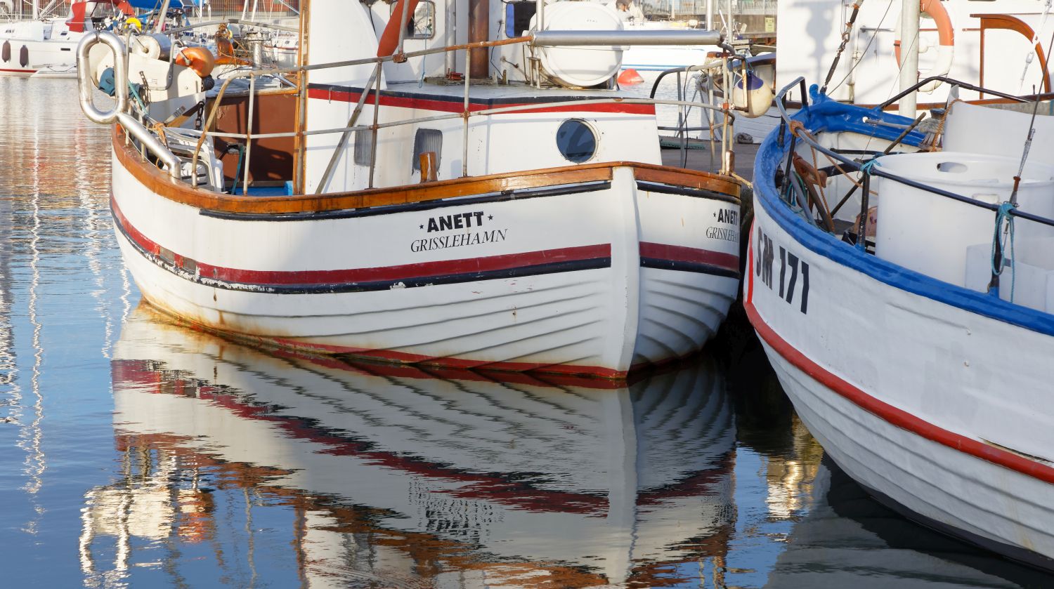 Fotografi föreställande en båthamn. En vitmålad, större båt i trä är i blickfånget. På båten står det Anett Grisslehamn med svarta bokstäver.