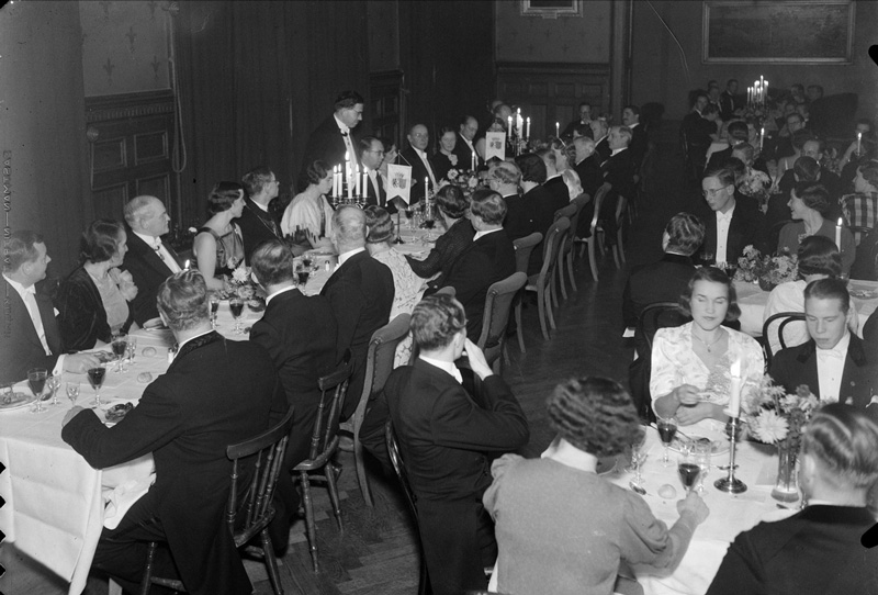 svartvitt fotografi med högtidsklädda människor sittande vid långbord.
