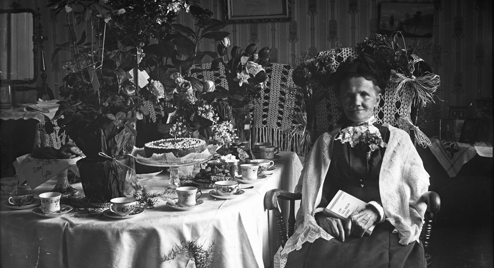 svartvitt fotografi föreställande kvinna som sitter vid ett bord uppdukat med fika och blommor