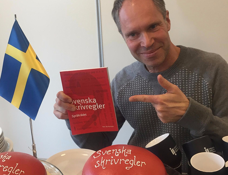 En man sitter vid ett bord dukat med kaffemuggar och två rosa tårtor med texten "Svenska skrivregler i glasyr". Det står även en svensk flagga på bordet, och mannen håller upp en röd liten bok som det också står Svenska skrivregler på. 