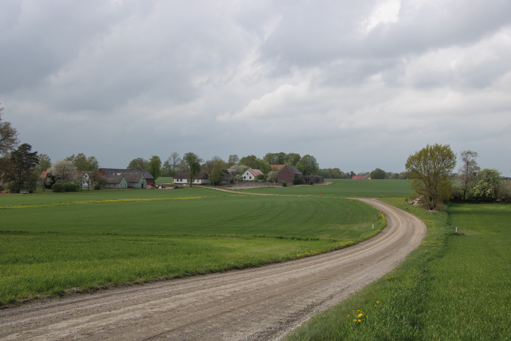 Fotot har i mitten en grusväg som leder fram till en liten by med några vita hus. Vägen kantas av gröna fält och i övre delen av fotot syns en mulen himmel.