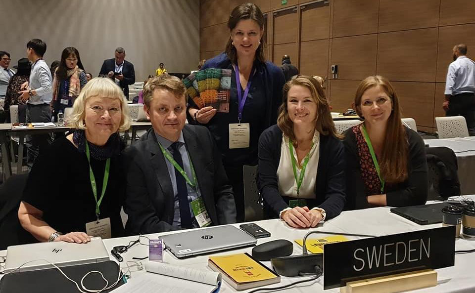 Bild från sammanträdeslokal, med Sveriges delegation vid ett bord som bär skylten "Sweden". 