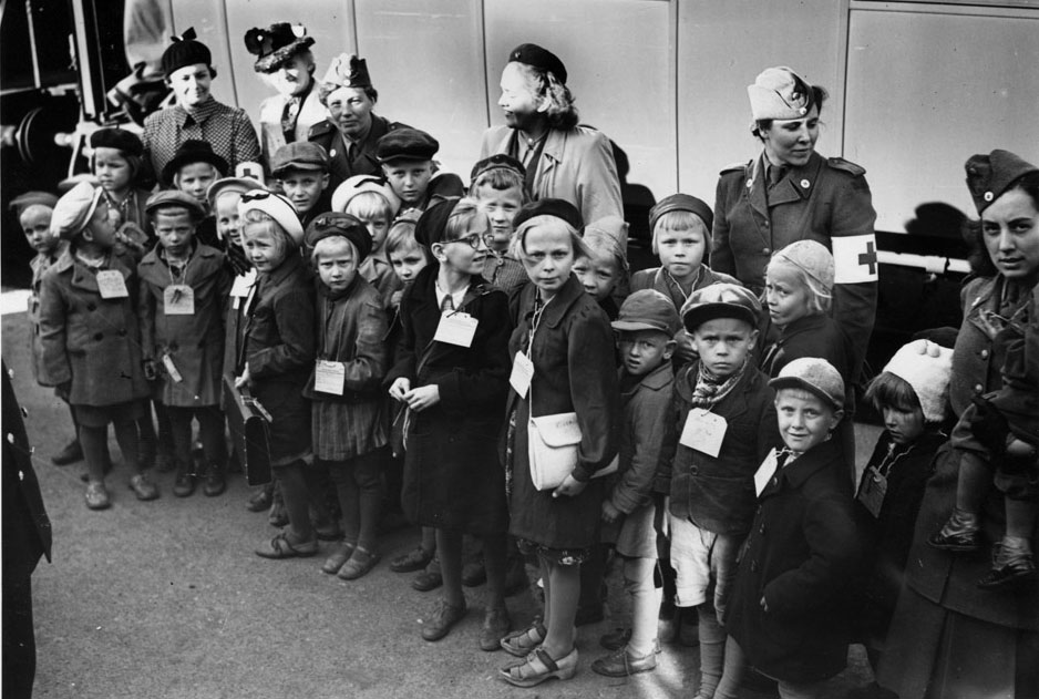 Svartvitt fotografi på en grupp barn med vinterkläder och namnlappar runt halsen.