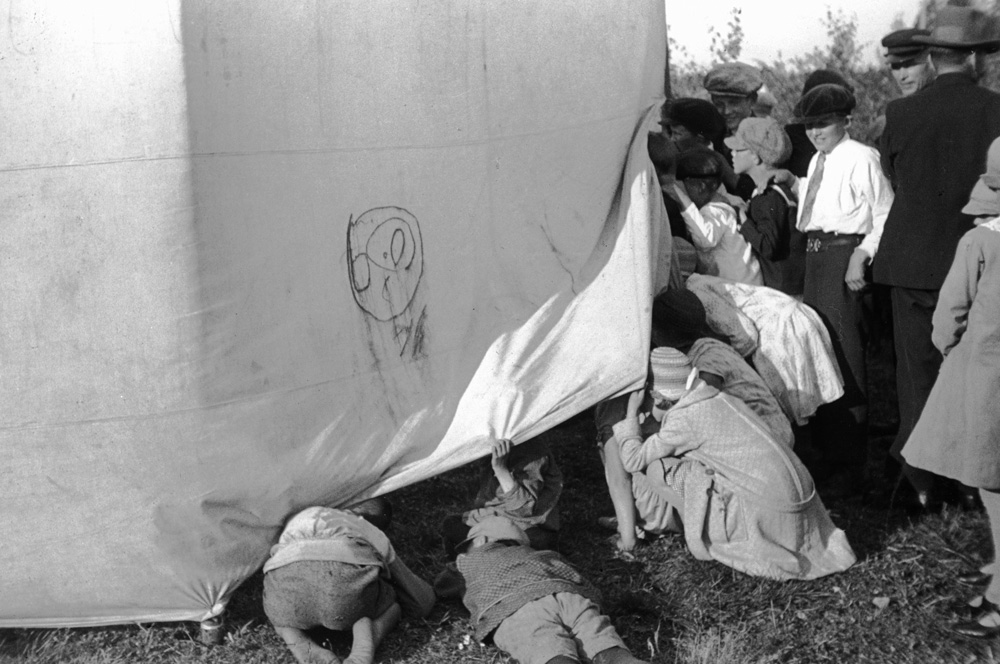 Svartvitt fotografi som föreställer flera barn som kikar in under en tältduk.