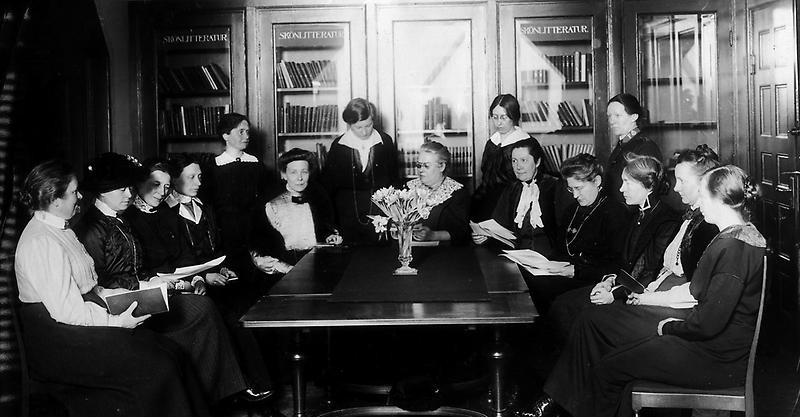 Grupp mörkklädda kvinnor som sitter runt ett bord i ett bibliotek.