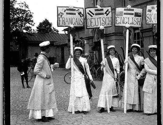 Kvinnor i vita långklänningar som håller stora skyltar med olika språk.