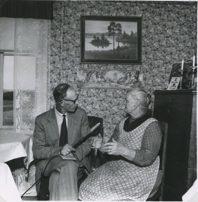 Svartvitt foto på en man som intervjuar en äldre kvinna. De sitter i ett rum på varsin stol och ser på varandra och samtalar. Han håller en stor mikrofon i handen. 
