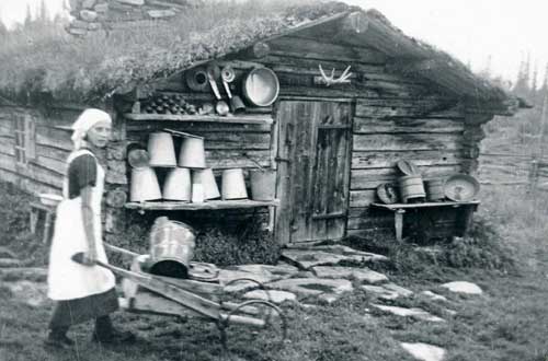 Kvinna drar skottkärra framför hus. 