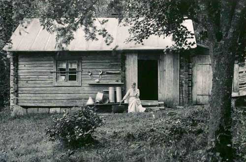 Kvinna sitter på farstubro utanför hus med gryta bredvid sig.