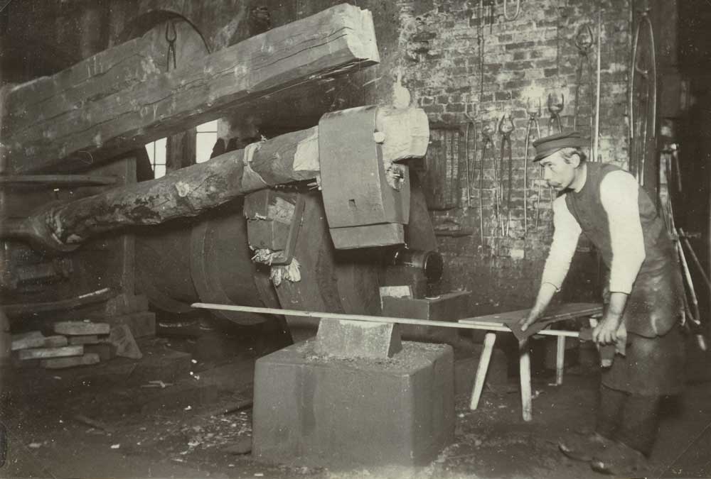 Svartvitt fotografi med man som arbetar vid en stor hammarliknande maskin. På väggen hänger verktyg.