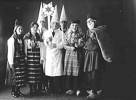 Svartvitt fotografi av en grupp ungdomar klädda som stjärngossar och i folkdräkt samt två tomtar med masker för ansiktena.