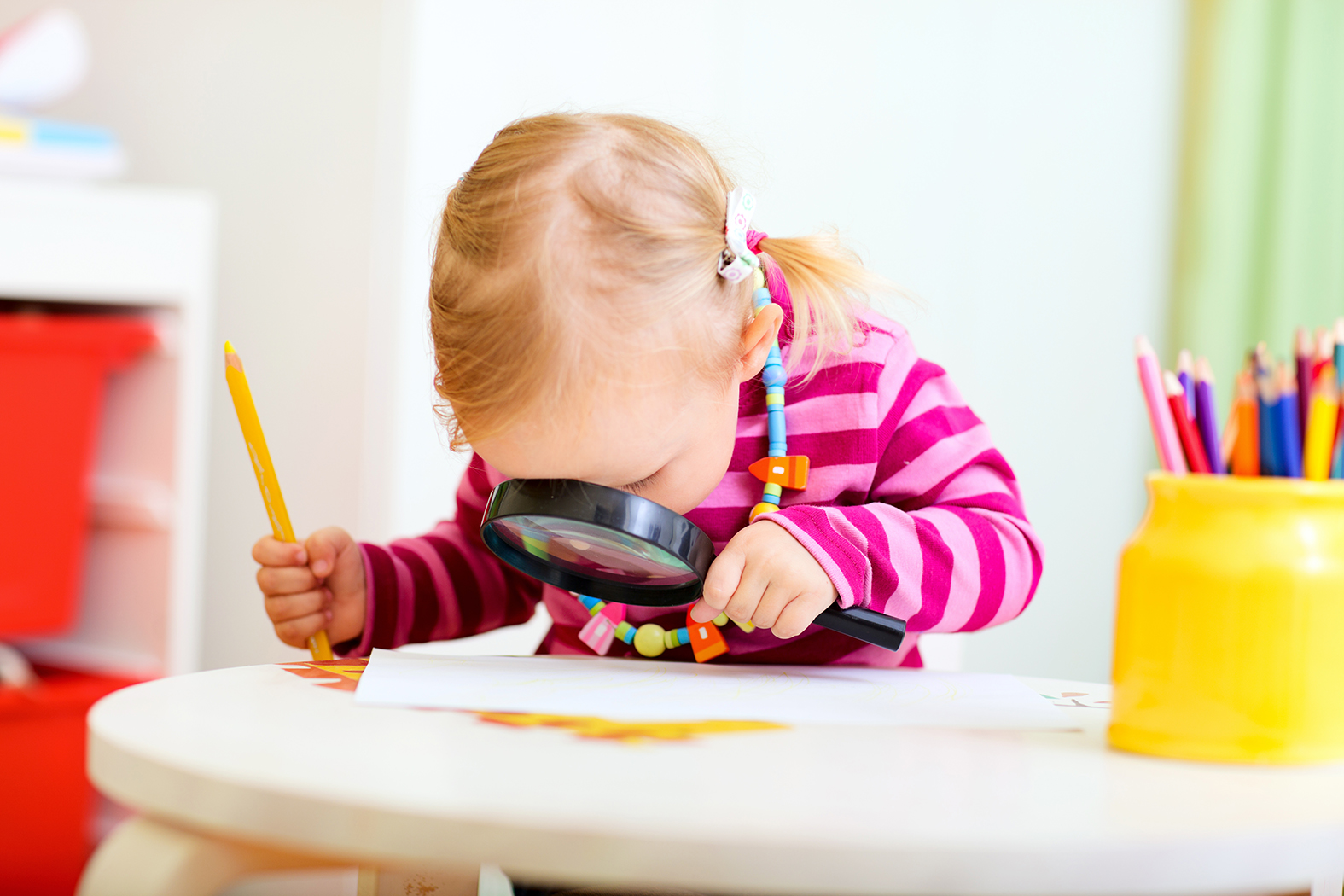 Ett förskolebarn i färgglad tröja sitter lutad över ett bord och tittar koncentrerat ner genom ett förstoringsglas.