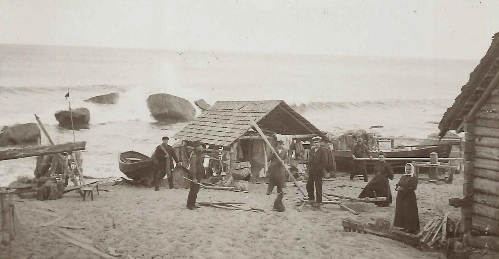 svartvitt fotografi med några människor som står mellan byggnader på en strand med havet i bakgrunden