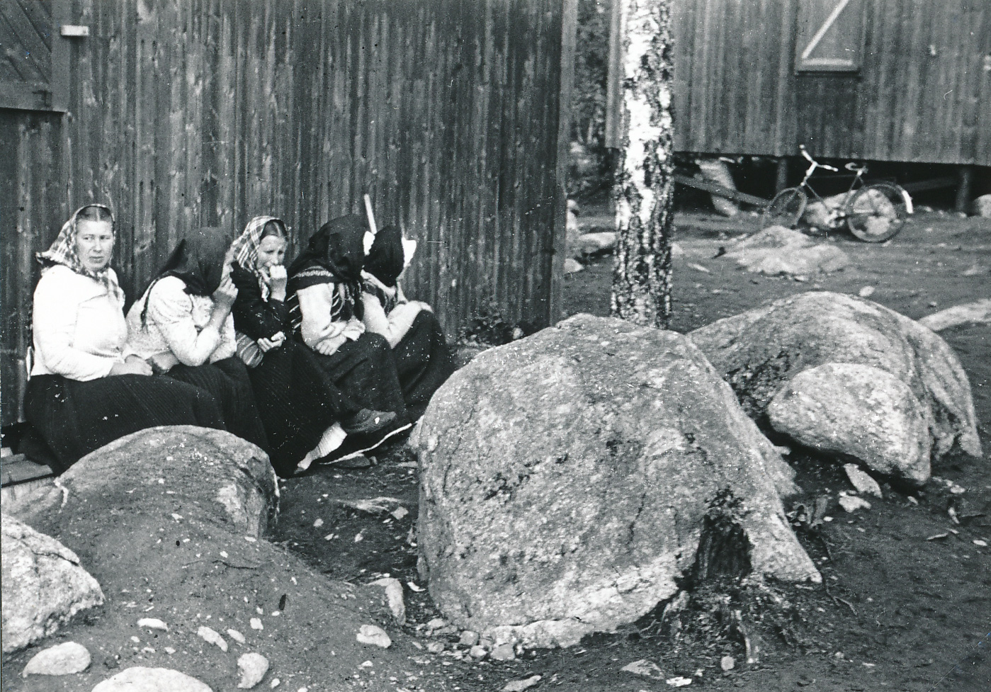 Svartvitt fotografi på fyra huckleklädda kvinnor som sitter mot en husvägg och tittar bortåt.