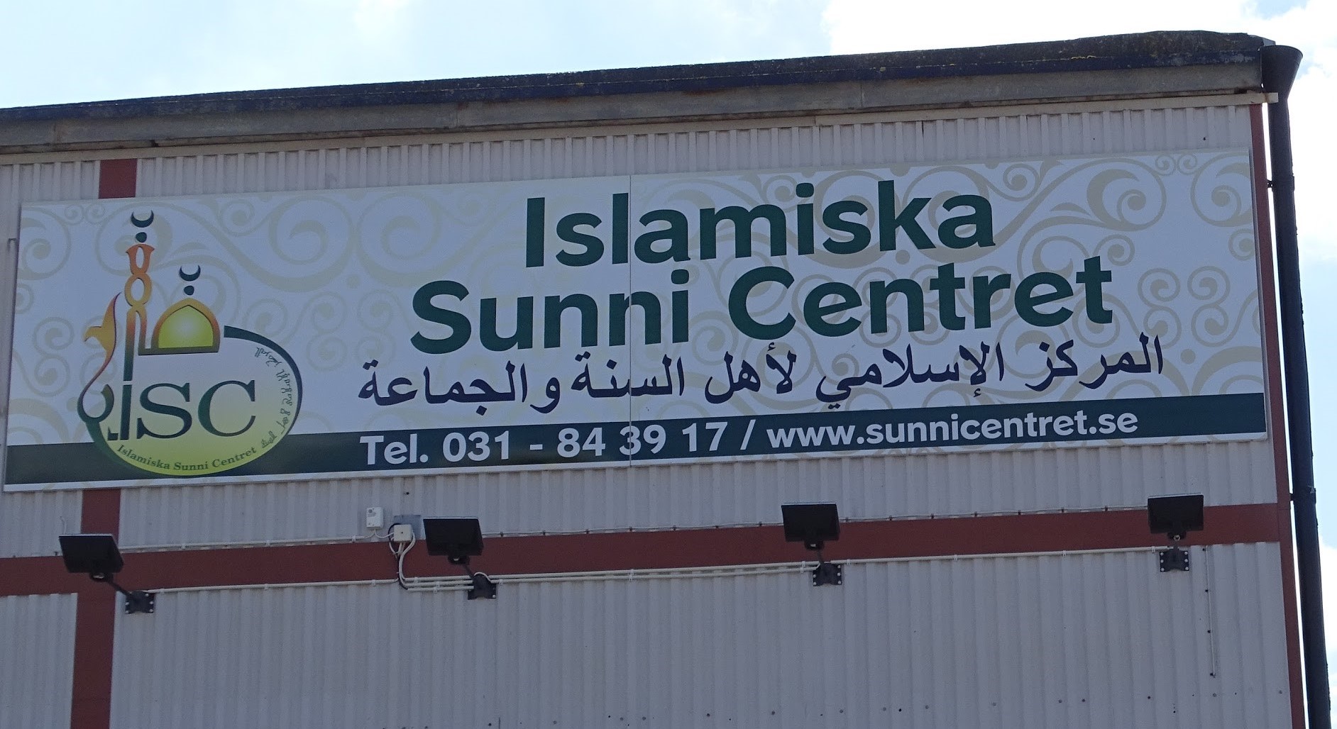 Bilden visar skylten till Islamiska Sunnicentret. Texten är grön och på skylten finns symboler kopplade till islam. Det svenska namnet står överst och det arabiska på raden under.