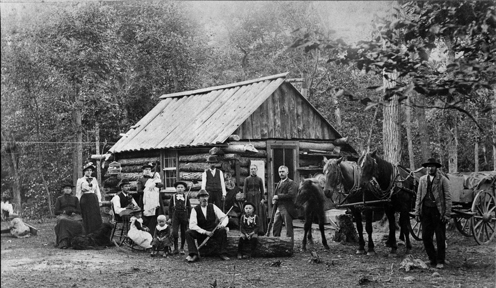 svartvitt fotografi med en familj och häst med vagn som står uppställda framför ett litet timrat hus.