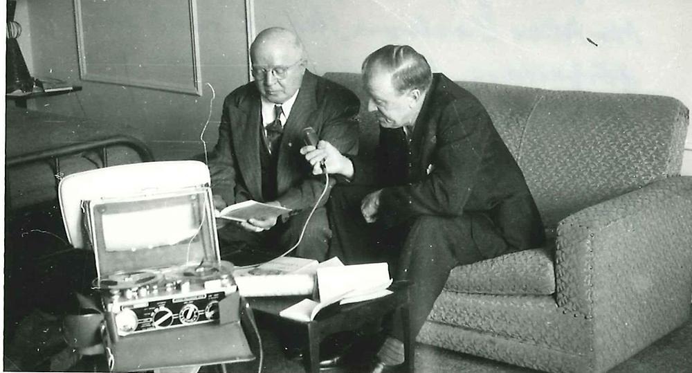 Svartvitt fotografi.Två män sitter på en soffa och pratar. Den ena mannen håller en mikrofon framför den andre och på bordet framför dem står en låda med inspelningsutrustning.