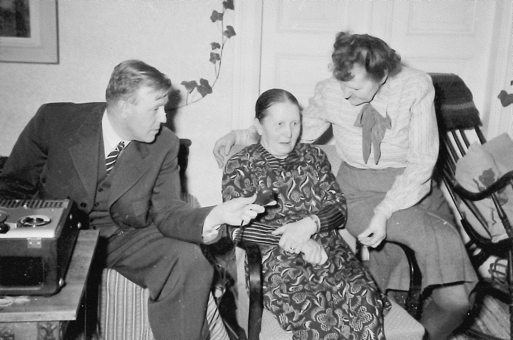 Svartvitt fotografi med en äldre kvinna som intervjuas av en kostymklädd man. Kvinnan sitter på en stol, och mannen sitter på stolskarmen och sträcker fram mikrofonen framför henne. På kvinnans andra sida sitter en yngre kvinna och tittar ner mot henne.