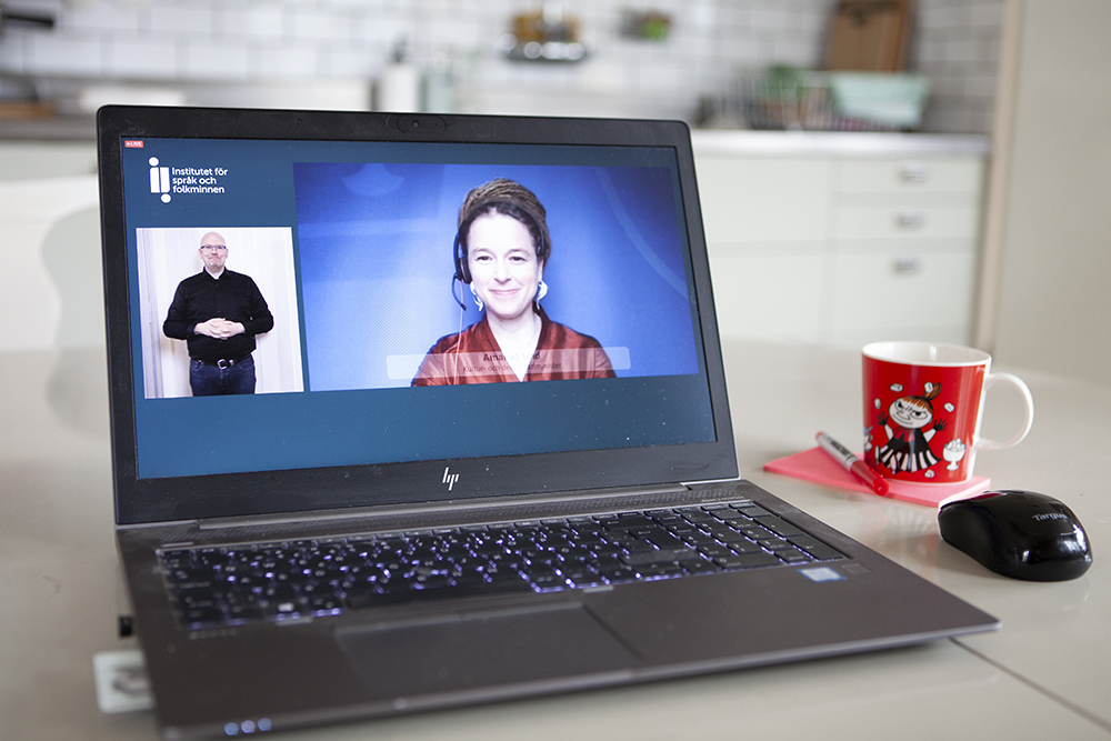 Foto på laptop på ett bord i ett kök. Även en mugg står på bordet. På skärmen syns ett pågående videomöte, med Amanda Lind och en teckenspråkstolk synliga i bild.