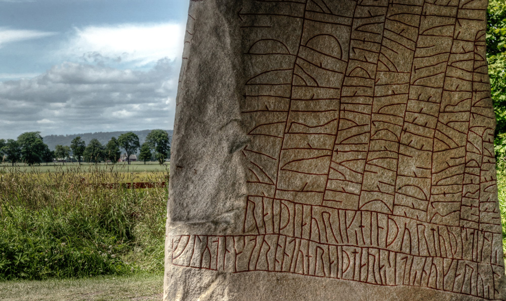 Närbild av runstenen Rökstenen där stenen med de rödmålade runorna syns i förgrunden och landskapet i bakgrunden.