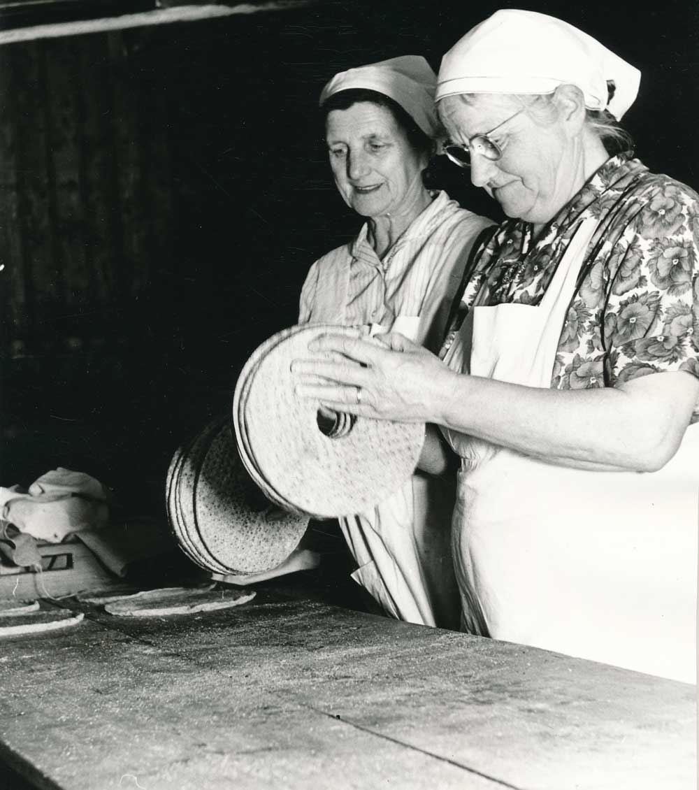 Två kvinnor står vid bakbord och formar brödkakor mellan händerna.