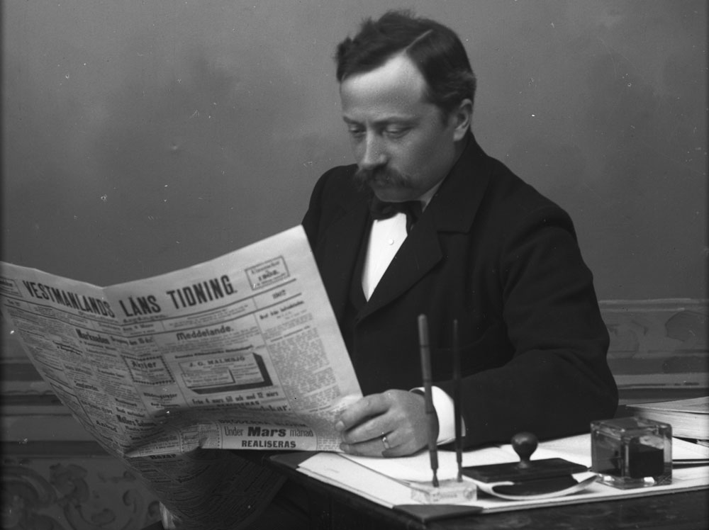 Svartvitt fotografi av man vid skrivbord som läser tidning.