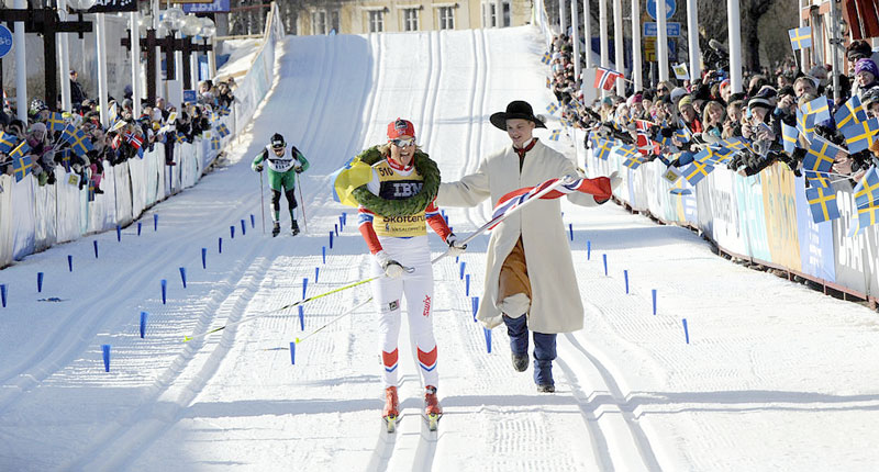 Man i  folkdräkt springer intill skidlöpare vid målgången. 