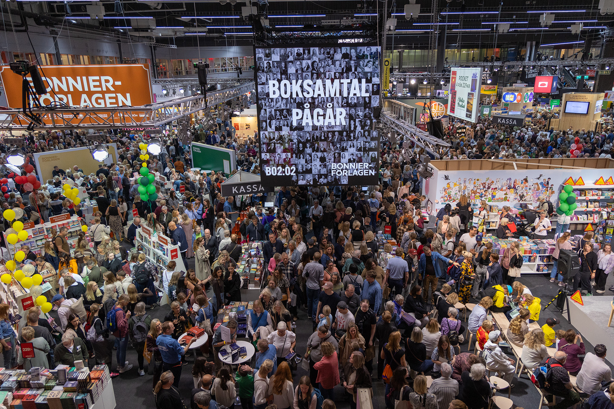 Vy ovanifrån över en stor mässhall fylld med människor som går runt bland montrar.
