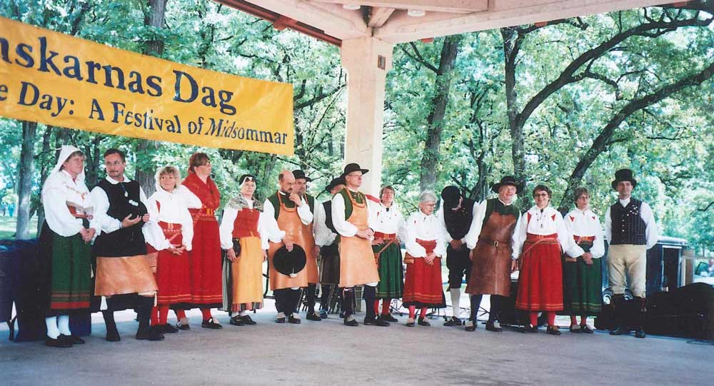 Människor i folkdräkt på scen under en paroll med texten Svenskarnas dag, A festival of Midsommar.