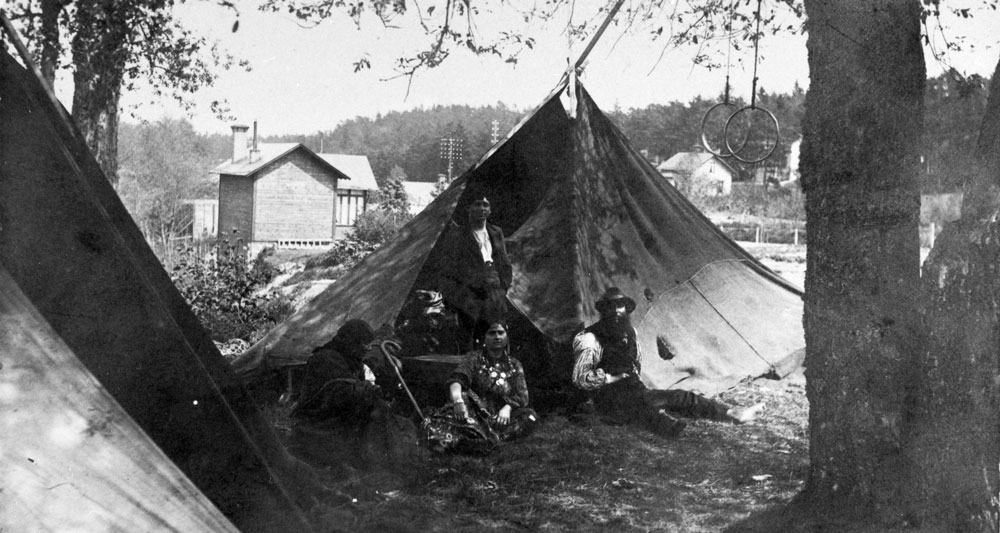 Svartvitt fotografi med personer som sitter framför ett tält i en träddunge.