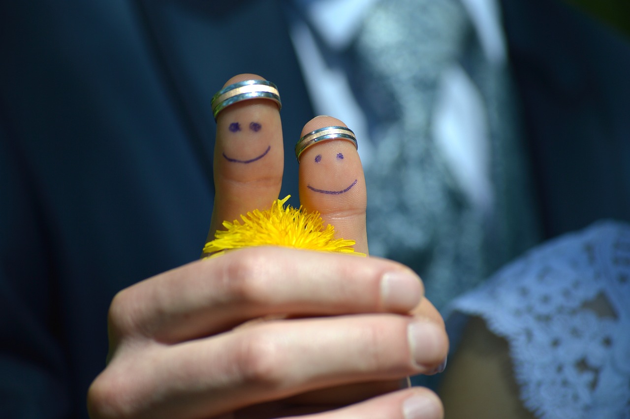 Foto föreställande ett handslag med två tummar som har målade glada ansikten och var sina förlovningsringar. I den hand som syns ligger en maskros.