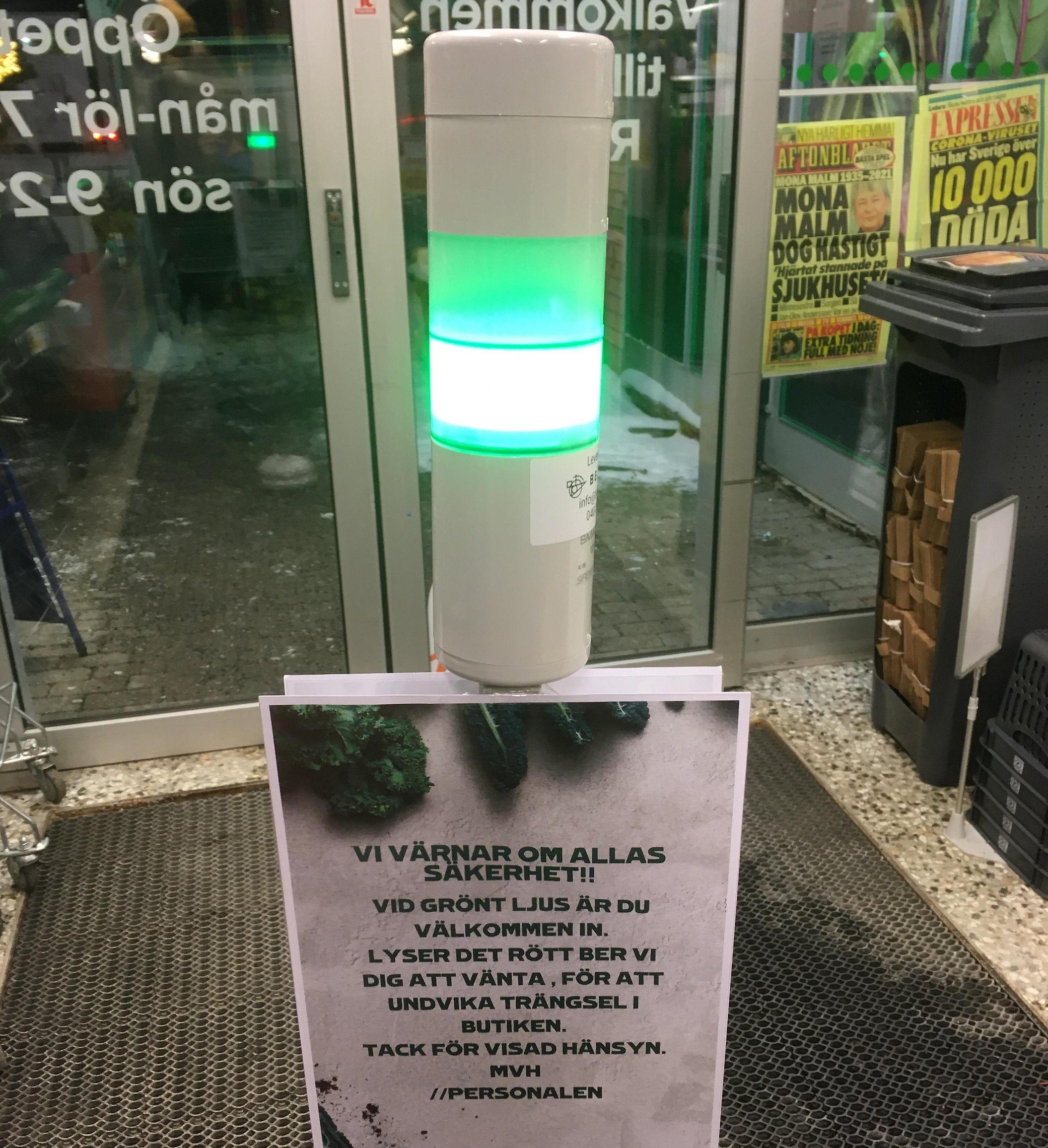 Fotografi på en grönt lysande lampa i ingången till en mataffär. En lapp
 sitter under, där det står "vi värnar om allas säkerhet! vid grönt ljus
 är du välkommen in. Lyser det rött ber vi dig att vänta, för att 
undvika trängsel i butiken. Tackför visad hänsyn. Mvh personalen".