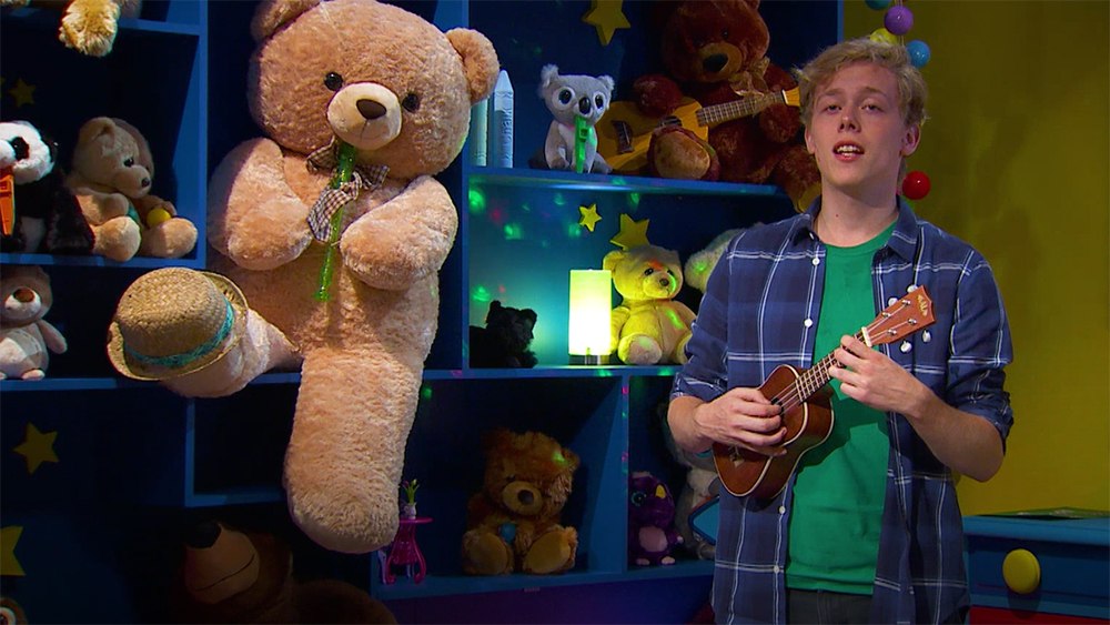 En ung person spelar på en leksaksgitarr i ett barnrum med en bokhylla full av leksaker.