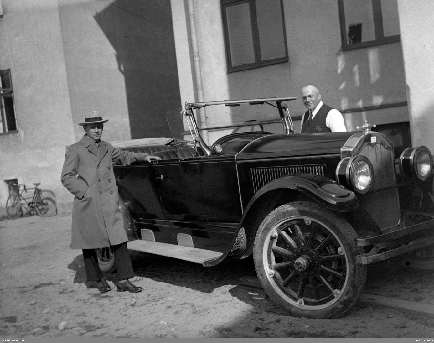 Två män poserar vid en parkerad äldre bil på en innergård, på en svartvit bild från 1926