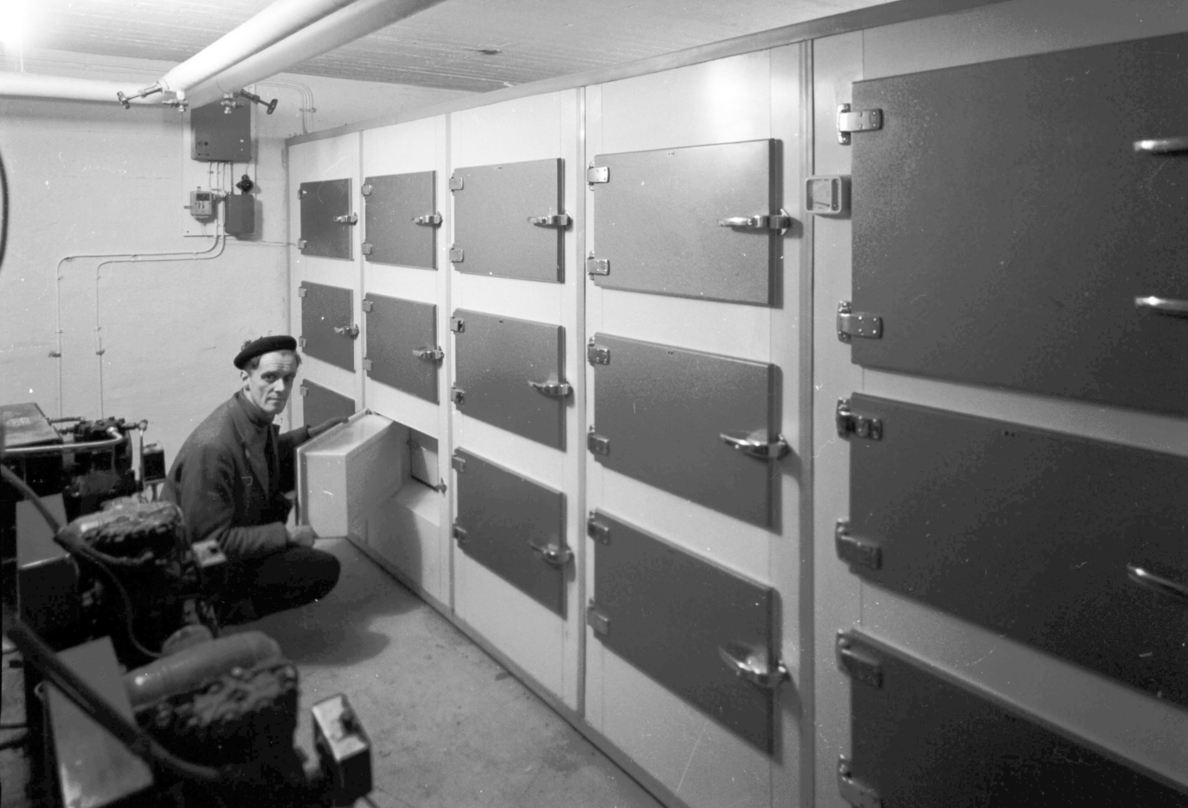 Svartvitt fotografi föreställande en vägg med luckor i rader. En man 
sitter på huk och ser in i kameran samtidigt som han öppnar en lucka på 
den understa raden. Dörren är tjock, ordentligt isolerad.