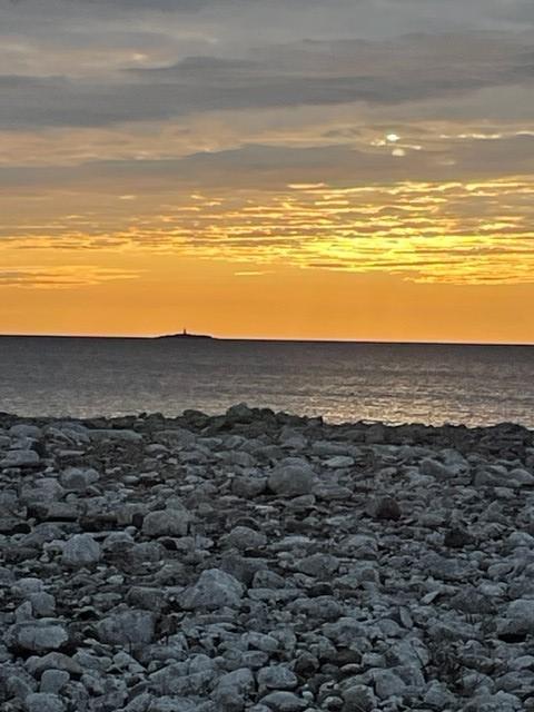 Foto från en stenig strand mot ett avlägset skär med fyr i solnedgång.