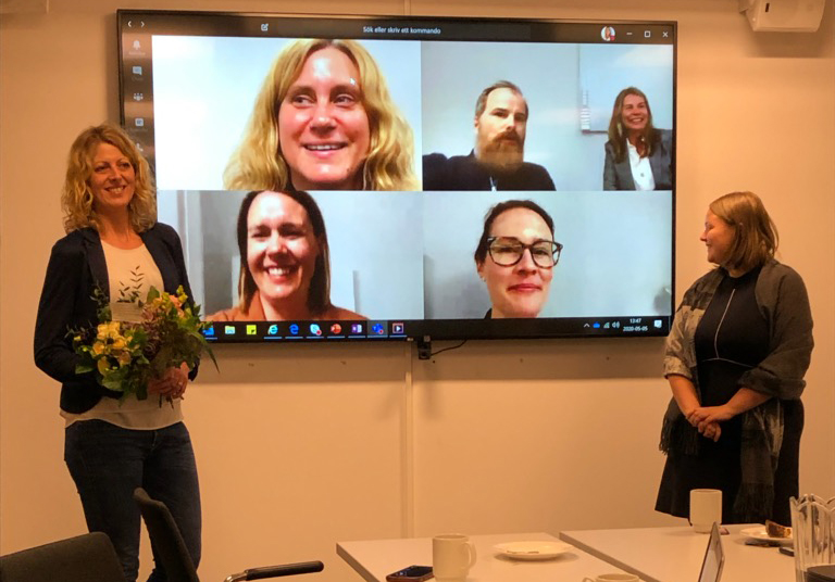 Fotografi föreställande två kvinnor som står i ett konferensrum framför en stor skärm. På skärmen syns ytterligare fem personer som deltar via länk. Alla ser glada ut. En av personerna framför skärmen har en bukett blommor i handen.