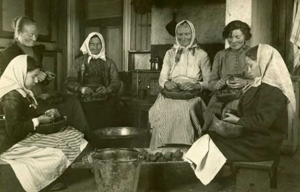 Svartvitt fotografi med kvinnor som sitter runt en balja potatis och skalar potatis.