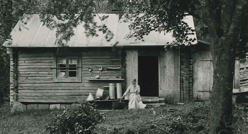 Kvinna som sitter på farstubron till ett mindre hus i trä.