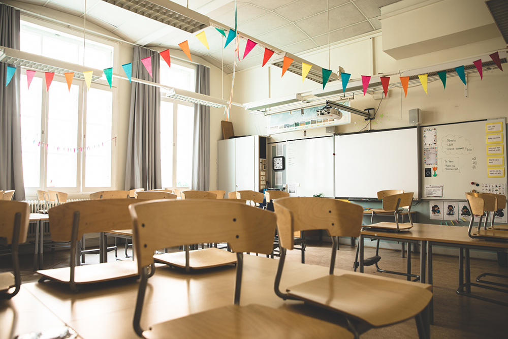 Fotografi på ett tomt klassrum med flera bordsgrupper. Stolarna är upphängda på borden. En färgglad vimpel hänger i taket.