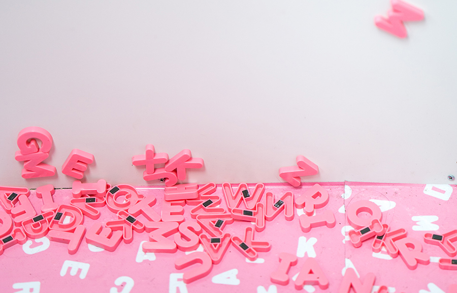 Bild på flertalet rosa kylskåpsmagneter föreställande bokstäver. Bokstäverna ligger huller om buller.