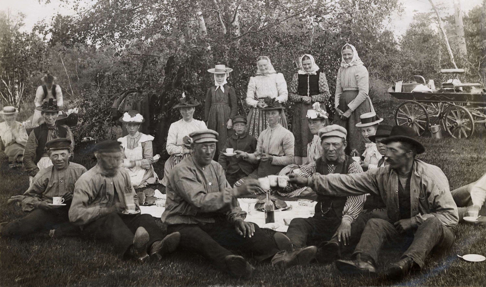 Svartvitt fotografi på en grupp människor som sitter och står i en gräsbacke. På marken är dukat med vit duk och bröd och de tre männen närmast kameran skålar med varandra. 