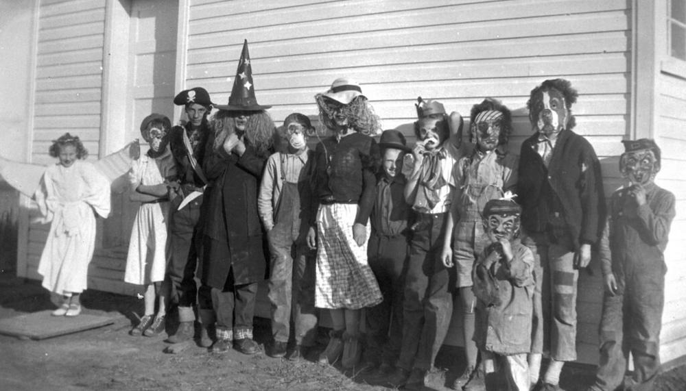 svartvitt fotografi av en grupp barn som står uppställda inför kameran framför ett hus. De är utklädda på olika sätt, till exempel till häxor och hundar. De flesta bär mask för ansiktet. 