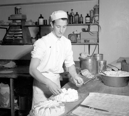 svartvitt fotografi med man i konditoriklädsel som står och gör i ordning en tårtliknande bakelse på ett bakbord